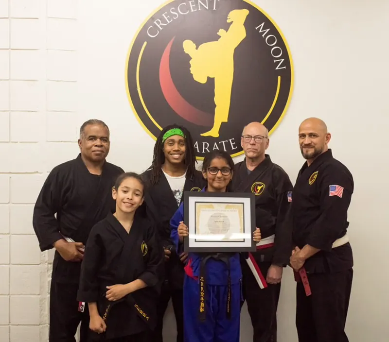 A student holds her framed rank certificate surrounded by her CMK instructors