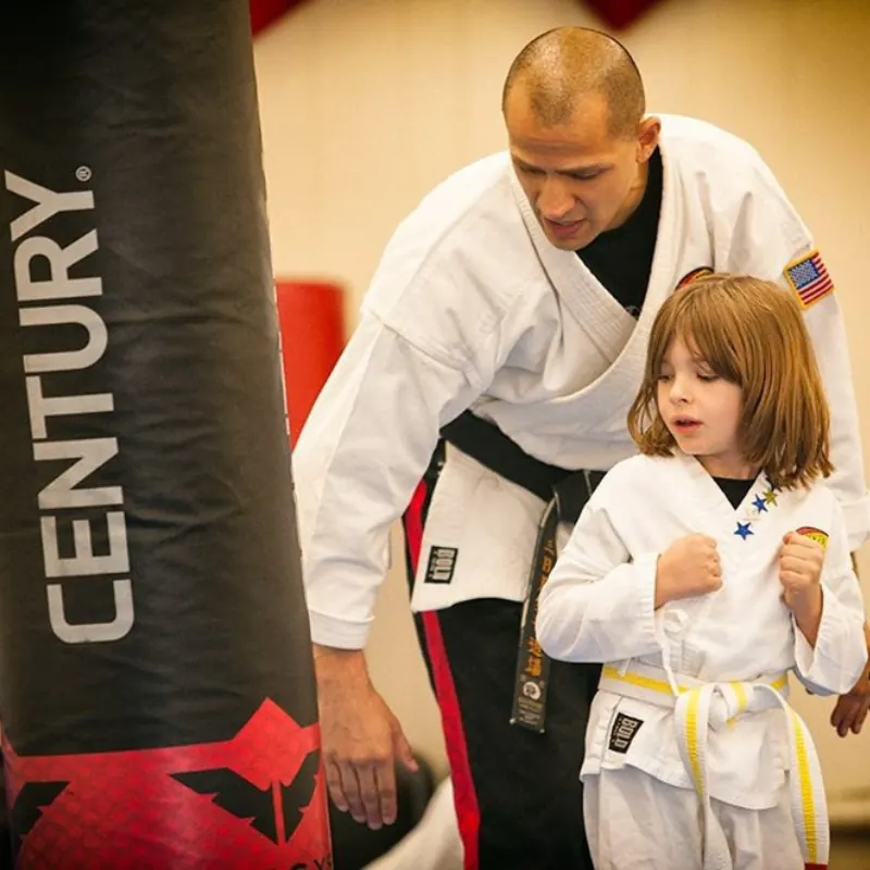 A black belt instructor kneels beside a young Little Ninjas student to guide her stance